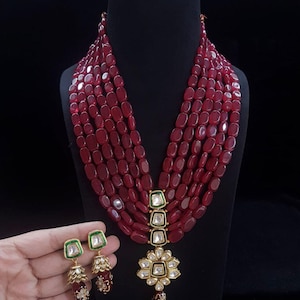May include: A maroon bead necklace and earring set. The necklace has multiple strands, a pendant with square and floral details, and matching earrings with green and gold accents. The jewellery is displayed on a black mannequin against a black background.