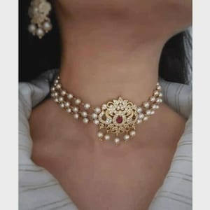 May include: A gold-toned choker necklace with multiple strands of small, round, white pearls. The centerpiece features an ornate, filigree design with clear and red gemstones. Matching pearl earrings are also visible.