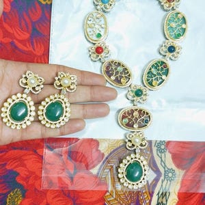 May include: A gold-toned necklace and matching earrings set. The necklace features oval pendants with colorful enamel designs and gemstone accents. The earrings have large green gemstone centers. The jewelry is displayed on a floral patterned fabric.