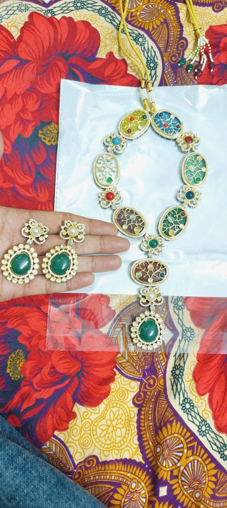 May include: A gold-toned necklace and matching earrings set. The necklace features oval pendants with colorful enamel designs and gemstone accents. The earrings have large green gemstone centers. The jewelry is displayed on a floral patterned fabric.