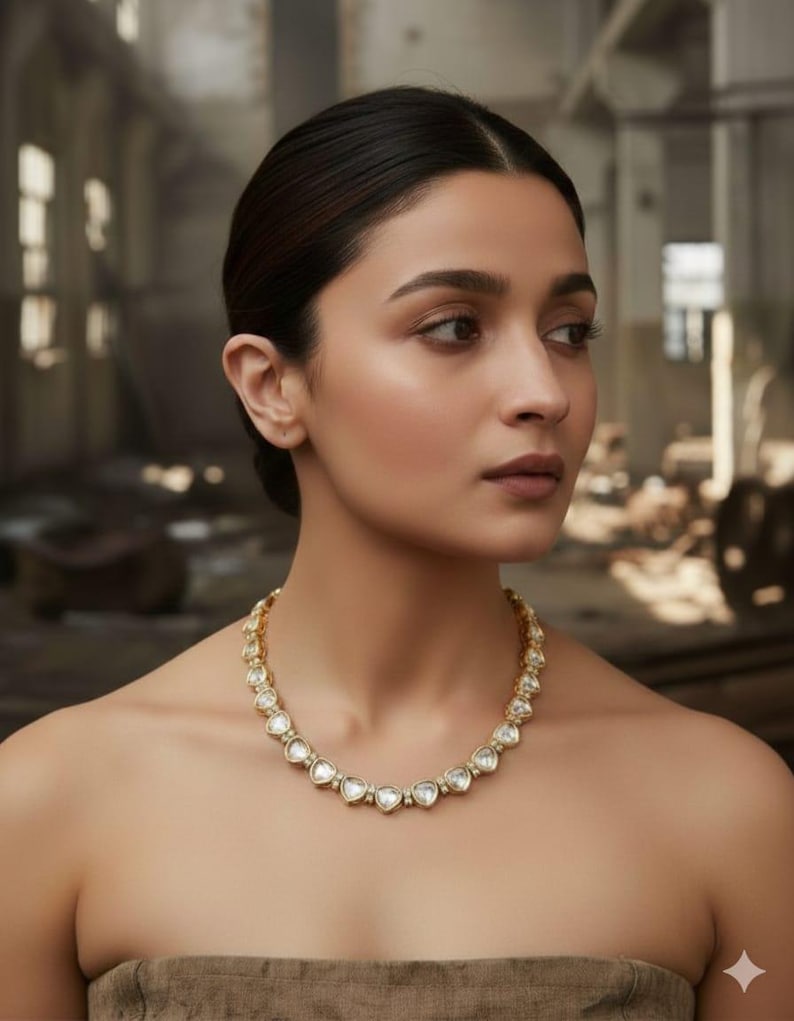 May include: A gold-toned necklace featuring heart-shaped clear stones. The necklace is worn around the neck. The background is blurred, with a neutral color palette. The necklace is the focal point of the image.