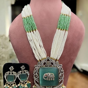 May include: An elaborate necklace and earring set. The necklace has multiple strands of white beads with green detailing, a large pendant with a green square centre, and an elephant design. The earrings have a similar design with green teardrop-shaped beads.