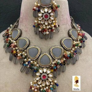 May include: A gray and gold-toned jewelry set featuring a necklace and a headpiece. The necklace and headpiece have gray teardrop-shaped stones, colorful beads, and floral designs with a central red stone. The set is displayed on a mannequin.