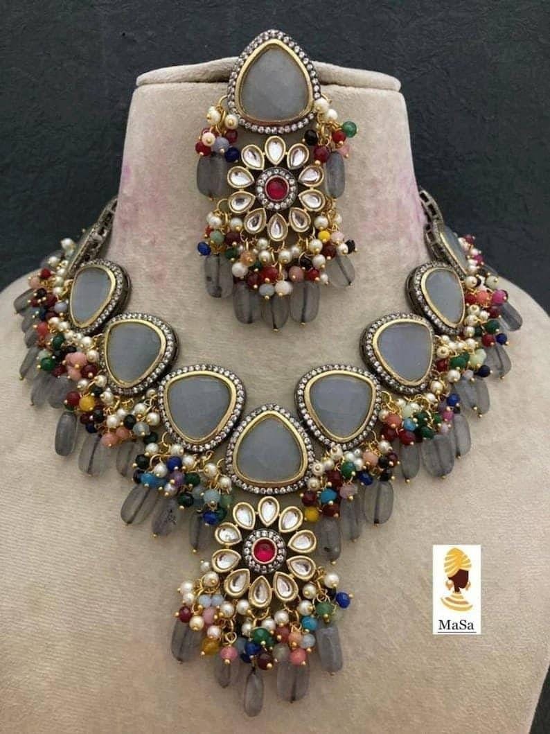 May include: A gray and gold-toned jewelry set featuring a necklace and a headpiece. The necklace and headpiece have gray teardrop-shaped stones, colorful beads, and floral designs with a central red stone. The set is displayed on a mannequin.