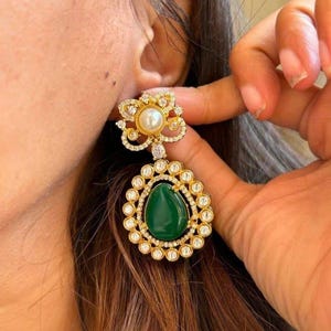May include: Gold-toned earrings with a floral design and a teardrop-shaped green stone. The earrings feature a pearl and small clear stones. The design is intricate and detailed, showcasing a classic jewelry style.