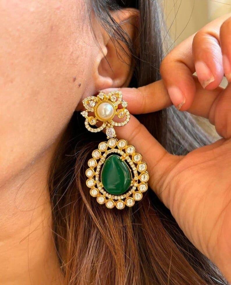 May include: Gold-toned earrings with a floral design and a teardrop-shaped green stone. The earrings feature a pearl and small clear stones. The design is intricate and detailed, showcasing a classic jewelry style.