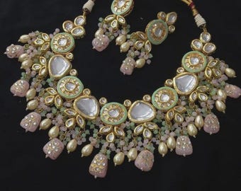 May include: A gold-toned necklace and earring set. The necklace features a central design with pale green and clear stones, surrounded by pink and white beads. The earrings mirror the necklace design. The jewelry is set against a black background.