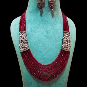 May include: A multi-strand red beaded necklace and matching earrings set. The necklace features ornate silver-toned clasps with floral and crystal accents. The earrings have a similar design with a dangling red bead. The jewelry is displayed on a turquoise mannequin.