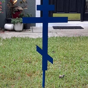 May include: A blue Orthodox cross stands in a grassy garden. The cross features a traditional three-bar design, with a top bar, a middle bar, and a slanted bottom bar. The cross is painted a solid, vibrant blue.