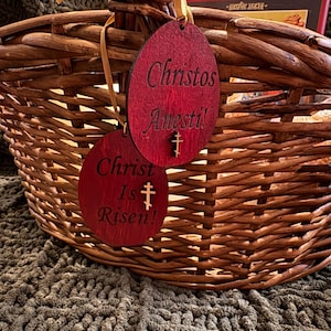 May include: A woven wicker basket with two red wooden egg-shaped ornaments. The ornaments have the phrases "Christos Anesti!" and "Christ Is Risen!" engraved on them, along with a cross symbol. The basket is tied with a gold ribbon.