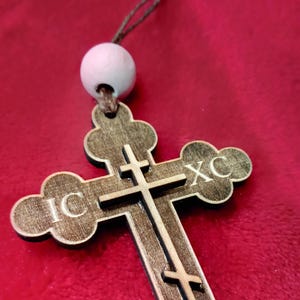 May include: A wooden cross with the letters "IC XC" engraved on it. The cross is brown and has a white bead at the top. The cross is hanging from a string.