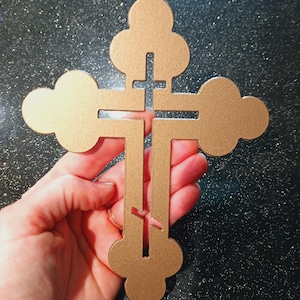 May include: A gold-coloured, ornate cross with a unique clover-like design on the ends of the arms. The cross is held in a hand against a dark, glittery background. The cross has a cutout design in the centre.
