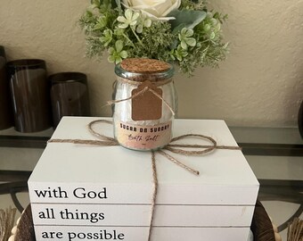 May include: A decorative arrangement featuring a glass jar of bath salt labeled "Sugar on Sunday", tied with twine, atop three white books with the text "with God all things are possible". A bouquet of white roses and greenery sits behind the jar.