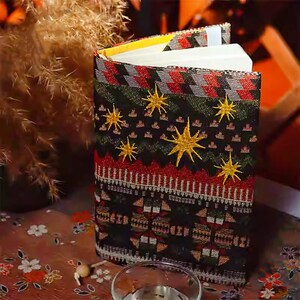 May include: A decorative journal with a patterned cover featuring gold star embroidery on a black background. The cover also has red, green, and white geometric designs. The journal's pages are visible, and the binding is yellow.