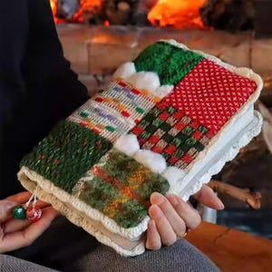 May include: A colorful, patchwork-style book cover with a white crocheted border. The cover features various textured fabrics in green, red, and white, with small pom-poms and decorative tassels. The overall theme is cozy and handcrafted.