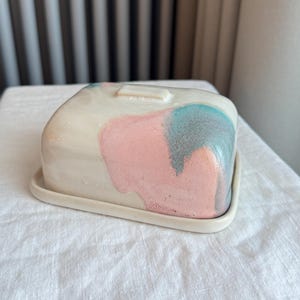 Può includere: Una burriera in ceramica con coperchio. Il piatto è rettangolare con bordi arrotondati e una base bianca. Il coperchio presenta macchie di vernice rosa e turchese. Il piatto poggia su una superficie bianca.