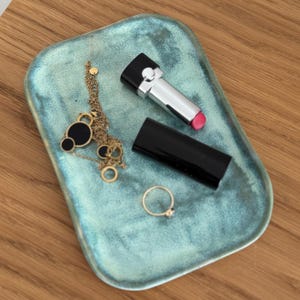 Könnte beinhalten: Ein tealfarbener Keramiktablett mit einer goldenen Kettenkette, einem schwarzen Lippenstift und einem schwarzen Eyeliner.
