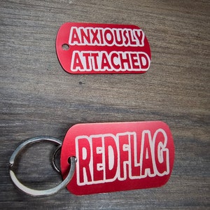 May include: Two red dog tag-shaped keychains with white text. One says "ANXIOUSLY ATTACHED" and the other says "REDFLAG". The keychain has a silver ring.