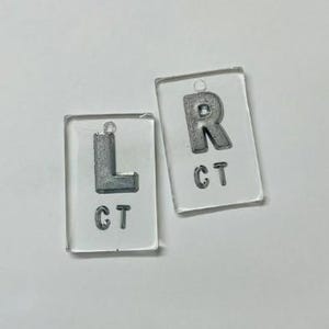 Puede incluir: Dos llaveros rectangulares transparentes con letras de metal plateado y las letras "CT". Un llavero tiene la letra "L", el otro la letra "R". Cada llavero tiene un pequeño agujero en la parte superior.