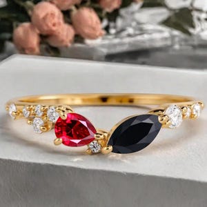 May include: A gold ring featuring a red pear-shaped gemstone and a black marquise-cut gemstone. The ring is adorned with small, round, clear gemstones. The ring's band is thin and gold-colored.