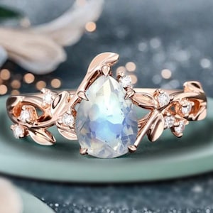 May include: Rose gold ring featuring a pear-shaped, iridescent moonstone. The band is adorned with delicate leaf and vine details, accented by small, sparkling gemstones. The ring sits on a light green surface, with a blurred background.