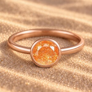Op de afbeelding: Een roségouden ring met een ronde, gefacetteerde oranje edelsteen in een gladde zetting. De steen heeft een levendige, sprankelende textuur met interne insluitingen en de ringband heeft een gladde, gepolijste afwerking met een warme kleur.