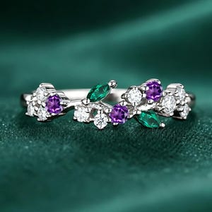 May include: A silver ring featuring a floral design with purple, green, and white gemstones. The ring has a delicate, nature-inspired aesthetic, with the gemstones arranged to resemble leaves and flowers. The ring is set against a dark green background.