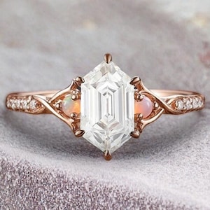 May include: A rose gold engagement ring with a hexagonal, emerald-cut center stone and two smaller, round stones on either side. The ring has a delicate, twisted band with small diamonds.