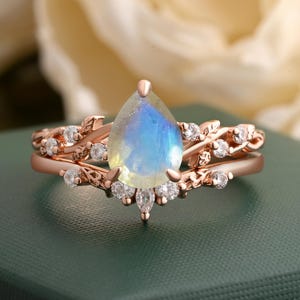 May include: Rose gold ring set with a pear-shaped, iridescent gemstone. The ring is adorned with small, round, and marquise-cut clear gemstones. The band has a delicate, leaf-like design. The ring is set on a green surface.