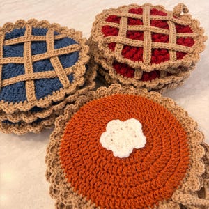 May include: A collection of round, crocheted pot holders designed to look like pies. The set includes blue and red berry pies with tan lattice crusts, and an orange pumpkin pie topped with a small, white, five-petaled crocheted flower.