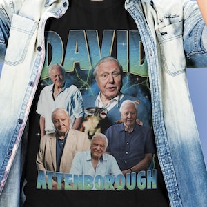May include: Black t-shirt with a graphic of David Attenborough and the text "DAVID ATTENBOROUGH" in blue and green. The graphic includes multiple images of Attenborough. The shirt is worn by a person.