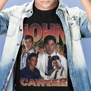 May include: Black t-shirt featuring a vintage-style graphic of four men in medical attire with the text "JOHN CARTER" in a retro font. The design includes a lightning bolt effect and a faded, aged aesthetic.