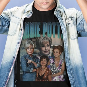 May include: A black short-sleeved t-shirt featuring a graphic print of the actress Annie Potts in various roles. The text "ANNIE POTTS" is printed in large, bold, teal-colored letters at the top of the graphic. The shirt is worn under a light blue denim shirt.