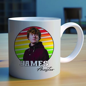 May include: White ceramic mug with a colorful retro-style graphic of a man with brown hair. The graphic includes a rainbow-colored sunburst design. The text "JAMES Acaster" is printed below the graphic.