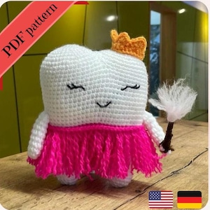 Peut inclure: Une poupée en crochet en forme de dent blanche portant une jupe rose avec une couronne dorée et tenant une baguette blanche et moelleuse. La poupée a des cils noirs et une bouche souriante.