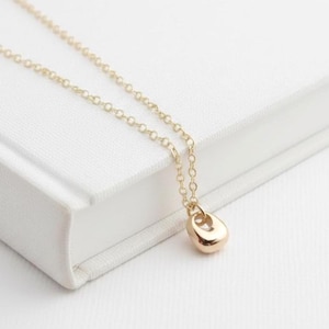 May include: A delicate gold-colored necklace with a teardrop pendant. The pendant has a smooth, polished surface and a small opening. The necklace chain is fine and also gold-colored. The jewelry is displayed on a white surface.