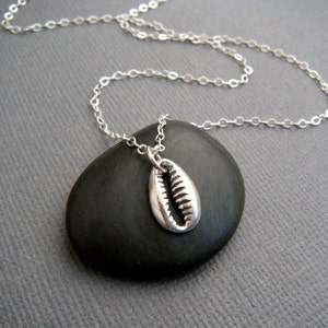 May include: A silver-colored cowrie shell pendant necklace. The pendant is a detailed replica of a cowrie shell, suspended from a delicate chain. The necklace is displayed on a dark gray surface.