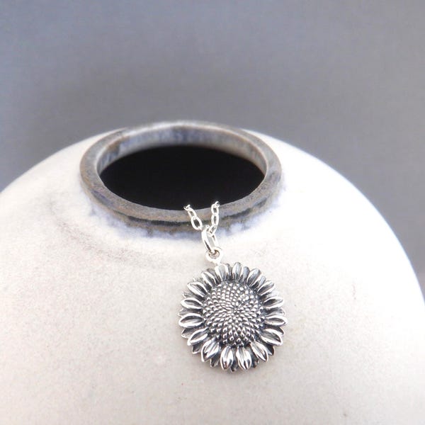 Può includere: Una collana con pendente a forma di girasole argentato. Il pendente è una rappresentazione dettagliata e tridimensionale di un girasole, con petali strutturati e un centro a capolino. La collana è esposta su uno sfondo neutro.