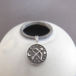 May include: Silver pendant necklace featuring a circular charm with a stylized depiction of a bow and arrow, a crescent moon, and a star. The charm has a textured, almost hammered, finish.