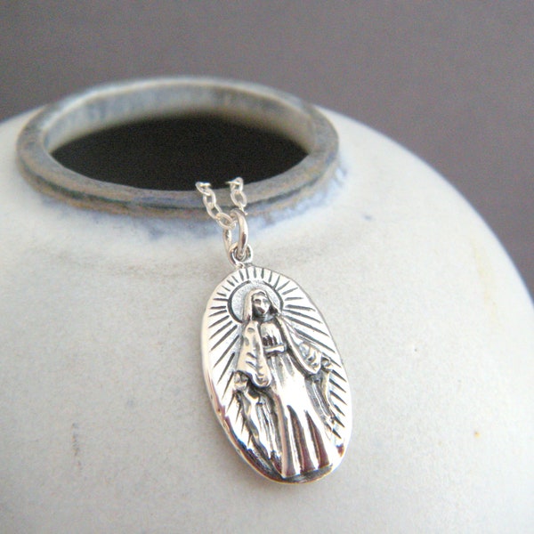 Può includere: Collana con ciondolo in argento con un'immagine dettagliata della Vergine Maria con un'aureola. Il ciondolo è di forma ovale e presenta un design testurizzato in rilievo.