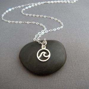 May include: A silver chain necklace with a small silver charm pendant featuring a wave design. The charm is circular and the wave is cut out of the metal.