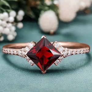 May include: Rose gold ring featuring a square, deep red gemstone center. The band is encrusted with small, clear gemstones. The ring is set against a teal background with blurred white flowers.
