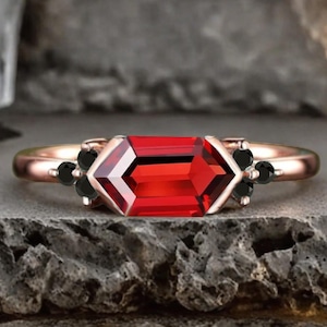 Peut inclure: Une bague en or rose ornée d'une grande pierre rouge rectangulaire et facettée. La bague est ornée de petites pierres noires de chaque côté de la pierre centrale. La bague est posée sur une surface grise texturée.