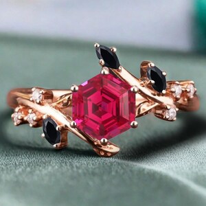 May include: Rose gold ring featuring a large, faceted, hexagonal ruby center stone. The ring is accented with black marquise-cut stones and small white diamonds, set on a branch-like design. The ring is displayed on a green surface.