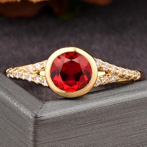Puede incluir: Un anillo de oro con una gran gema roja brillante y redonda. La gema está engastada en un bisel de oro, y la banda está adornada con pequeñas piedras blancas brillantes. El anillo se muestra sobre una superficie gris.