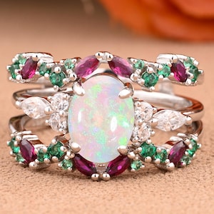 May include: A three-piece ring set featuring a large oval opal center stone. The rings are adorned with smaller gemstones in shades of green, red, and white, set in a silver band. The design is intricate and elegant, suitable for special occasions.