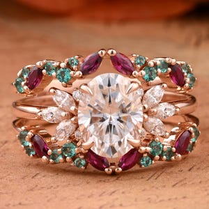 Peut inclure: Une bague de fiançailles en trois parties avec une grande pierre centrale ronde et claire entourée de pierres plus petites et claires et un halo de pierres en forme de marquise. La bande est ornée d'une rangée de pierres précieuses rouges et vertes.