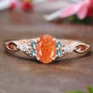 May include: A rose gold ring with an oval-shaped sunstone center stone surrounded by smaller green and white stones.