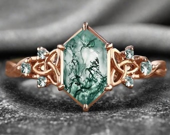 May include: Rose gold ring featuring a large, hexagonal green moss agate gemstone. The ring has intricate Celtic knot designs on the sides, accented with small, light blue gemstones. The band has a twisted rope design.
