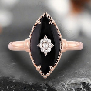 May include: A rose gold ring with a black onyx marquise-shaped stone set in a delicate, ornate design. The stone is accented with a small cluster of diamonds in the center.
